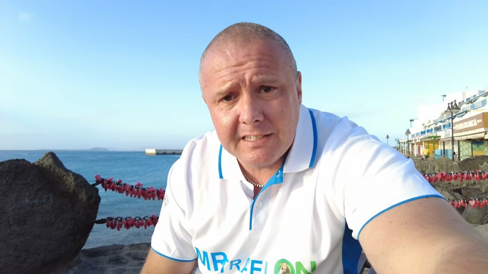 A man filming himself at a beach with love locks in the background.