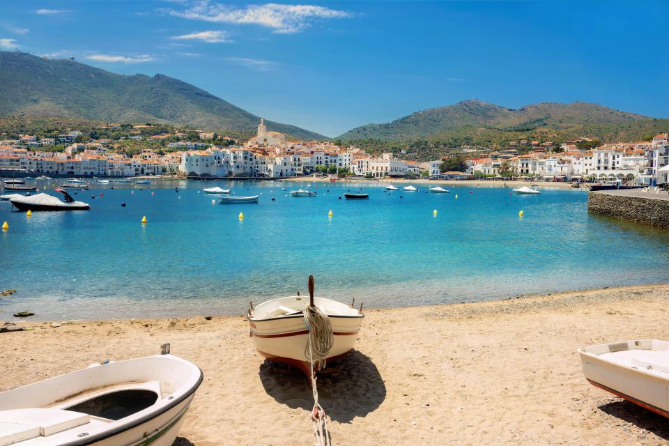 Cadaques, Costa Brava, Spain, with the town and boats on the water and a boat on the sandy shore.