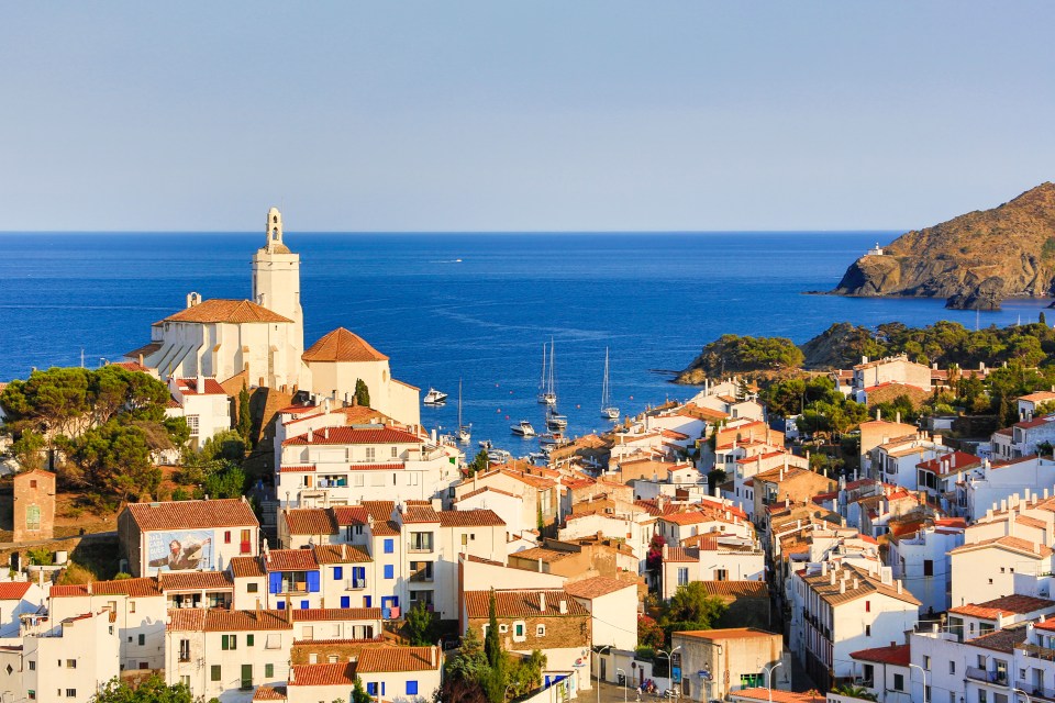 A hillside town with white buildings and terracotta roofs, overlooking a bay with boats, with a church steeple on the left and a lighthouse on a distant headland on the right.