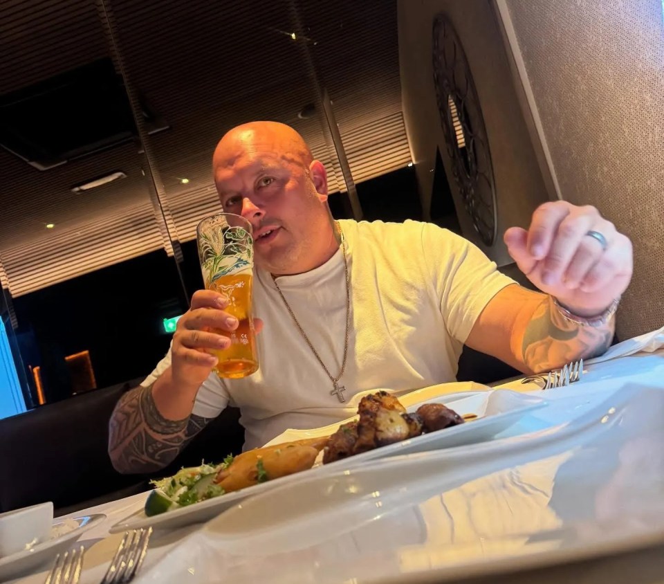 Bald man with tattoos on both arms holding a beer at a dining table.