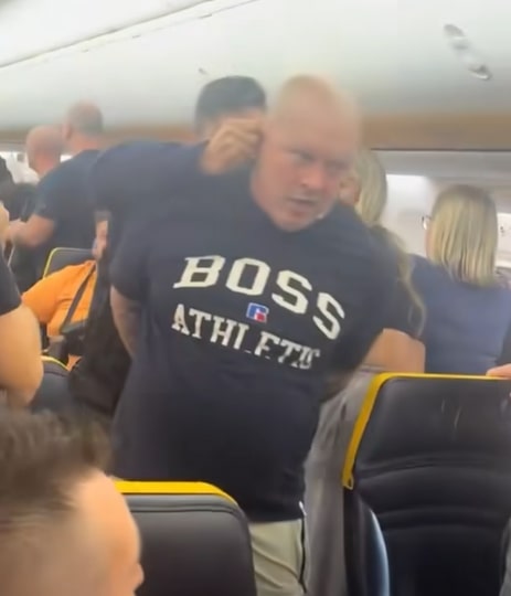 A drunk man in a "BOSS ATHLETIC" t-shirt being escorted off a Ryanair flight.