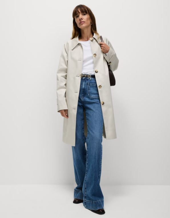 Woman wearing a cream raincoat, white t-shirt, blue wide-leg jeans, black belt, and brown handbag.