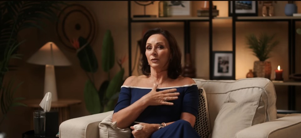 A woman with dark hair in a blue dress sits on a couch, holding her chest with one hand.