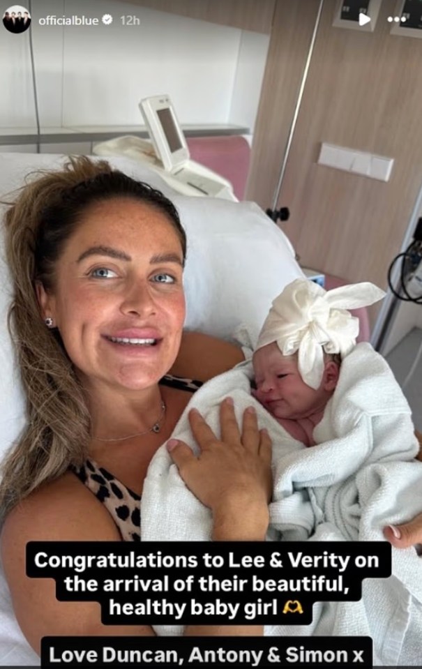 Woman smiling in a hospital bed holding a newborn baby wrapped in a white blanket and head covering.