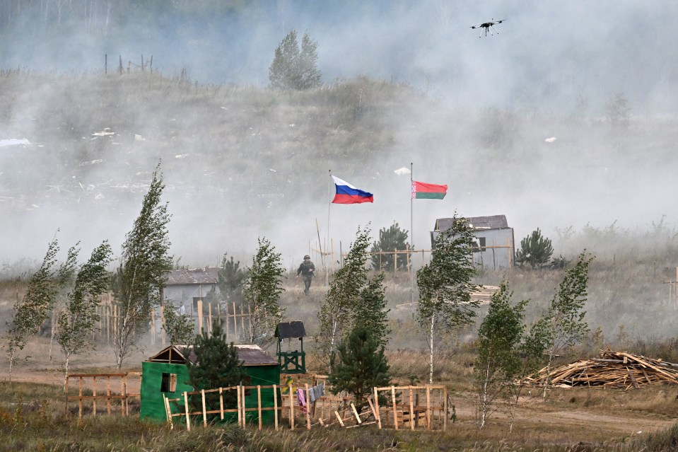 A drone flies over Russian and Belarusian flags during joint military drills, with a serviceman walking in the background amidst smoke and small buildings.