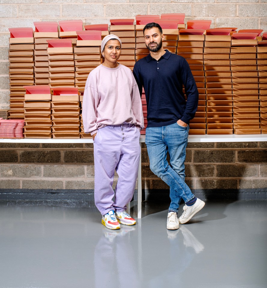 Zara and Zak in a warehouse, standing in front of stacks of pink and brown boxes.