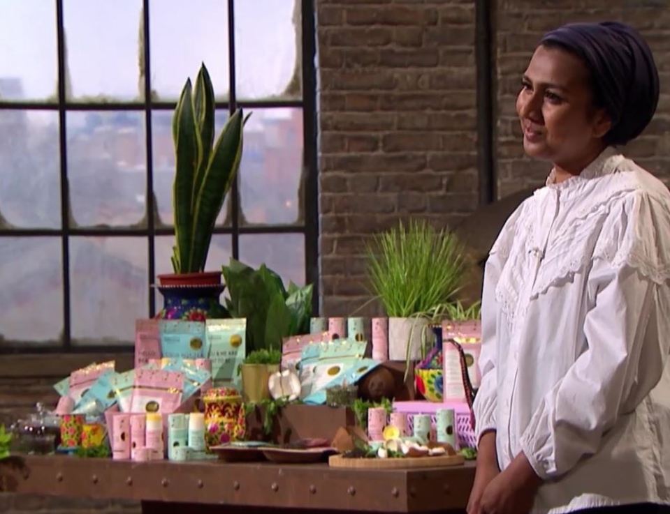 A woman in a white blouse and blue headscarf stands next to a table displaying various packaged products and plants.