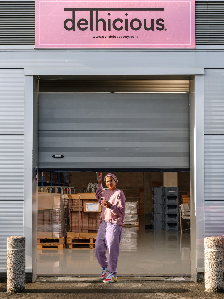 A person in purple outfit and headscarf holding a phone while standing in front of a warehouse entrance with "delhicious" sign above.