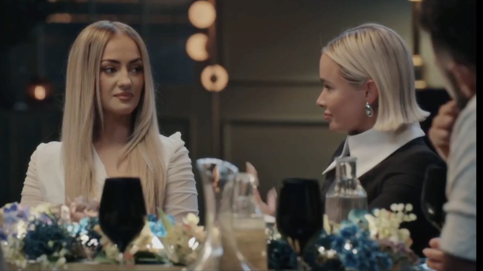 Two women at a dinner party, with the woman on the left wearing a white blazer and the woman on the right wearing a black dress with a white collar.