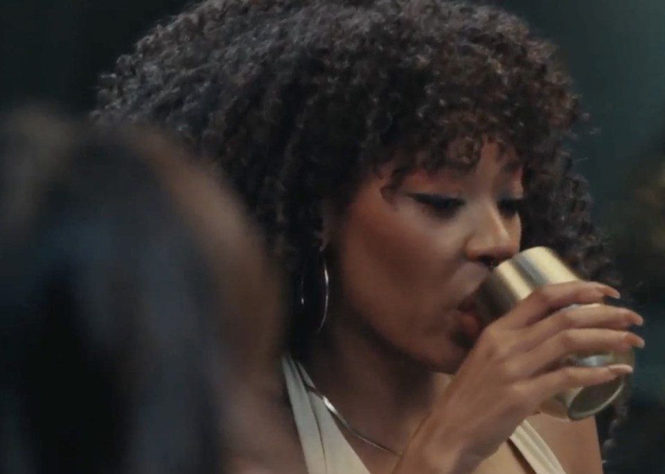 A person with curly dark hair and eyeliner drinking from a metal cup.