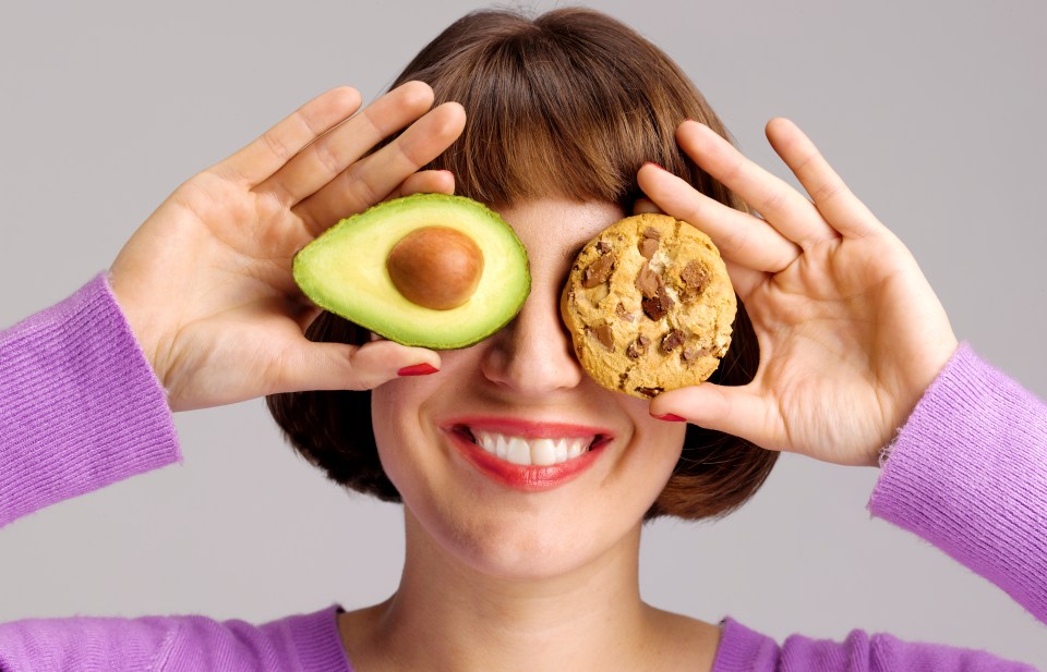 Woman holding an avocado slice over one eye and a chocolate chip cookie over the other, smiling.