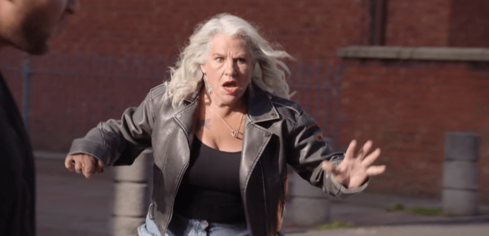 A woman with white hair, wearing a black leather jacket, yells at a man.
