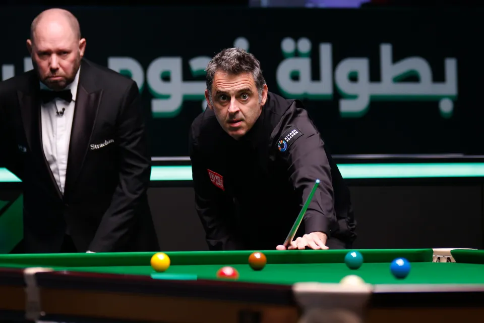 Ronnie O'Sullivan plays a shot in a snooker match with a referee in the background.