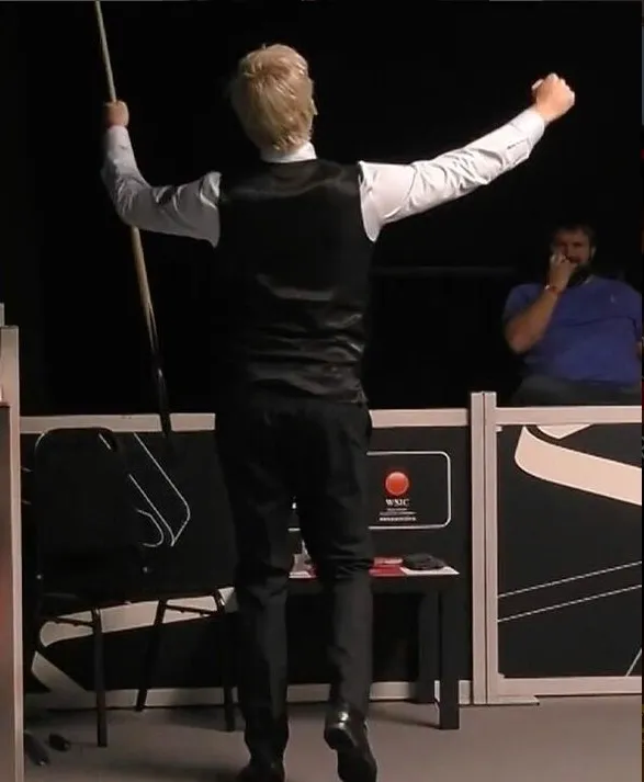A snooker player in a black vest and white shirt celebrates a shot, holding a cue stick.