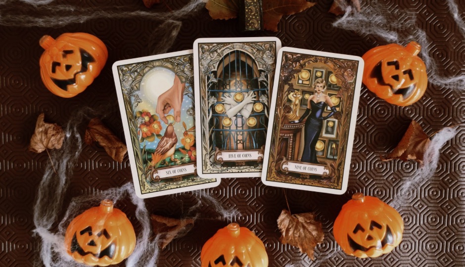 Three tarot cards, Six of Coins, Five of Coins, and Nine of Coins, are surrounded by Halloween decorations including pumpkins, spiderwebs, and leaves.