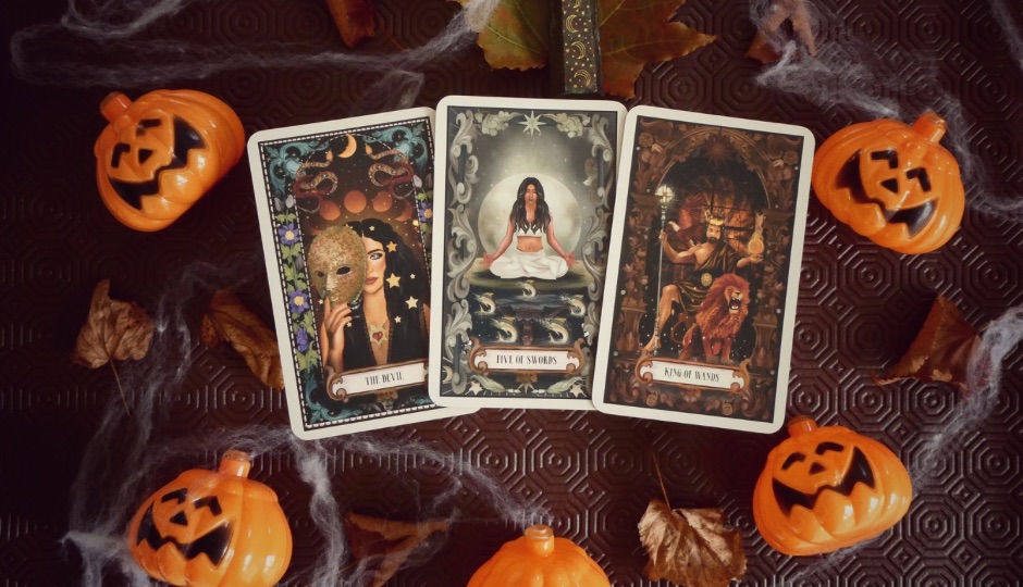 Three tarot cards: The Devil, Five of Swords, and King of Wands, surrounded by Halloween decorations.