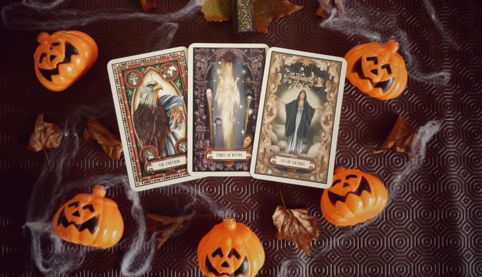 Three tarot cards "The Emperor", "Three of Wands", and "Ace of Swords" surrounded by Halloween decorations.