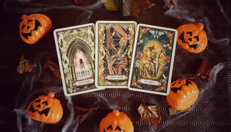 Three tarot cards (Six of Swords, Eight of Swords, and The Empress) surrounded by Halloween decorations.