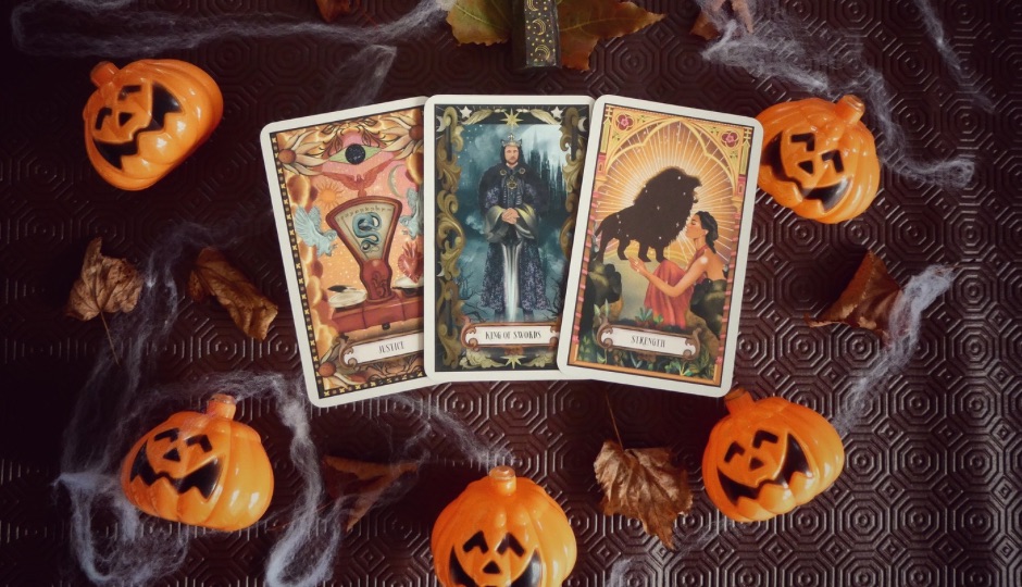 Three tarot cards, "Justice," "King of Swords," and "Strength," surrounded by decorative pumpkins, leaves, and cobwebs.