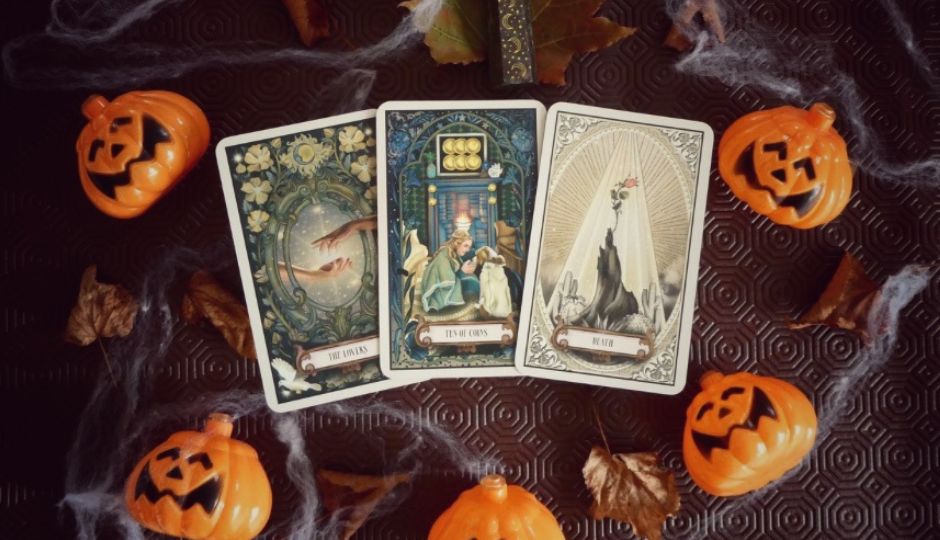 Tarot cards "The Lovers", "Ten of Cups", and "Death" surrounded by small pumpkin decorations, autumn leaves, and artificial spiderwebs.