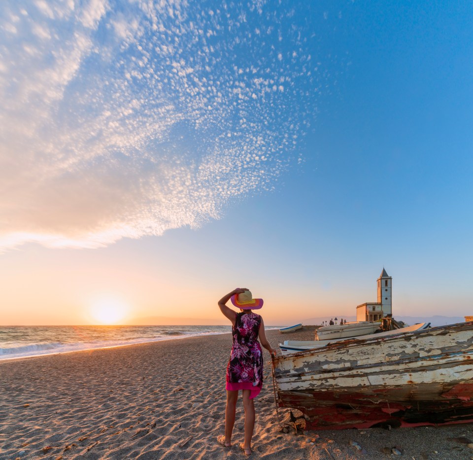 Woman looking at sunset over beach with old boat and church.