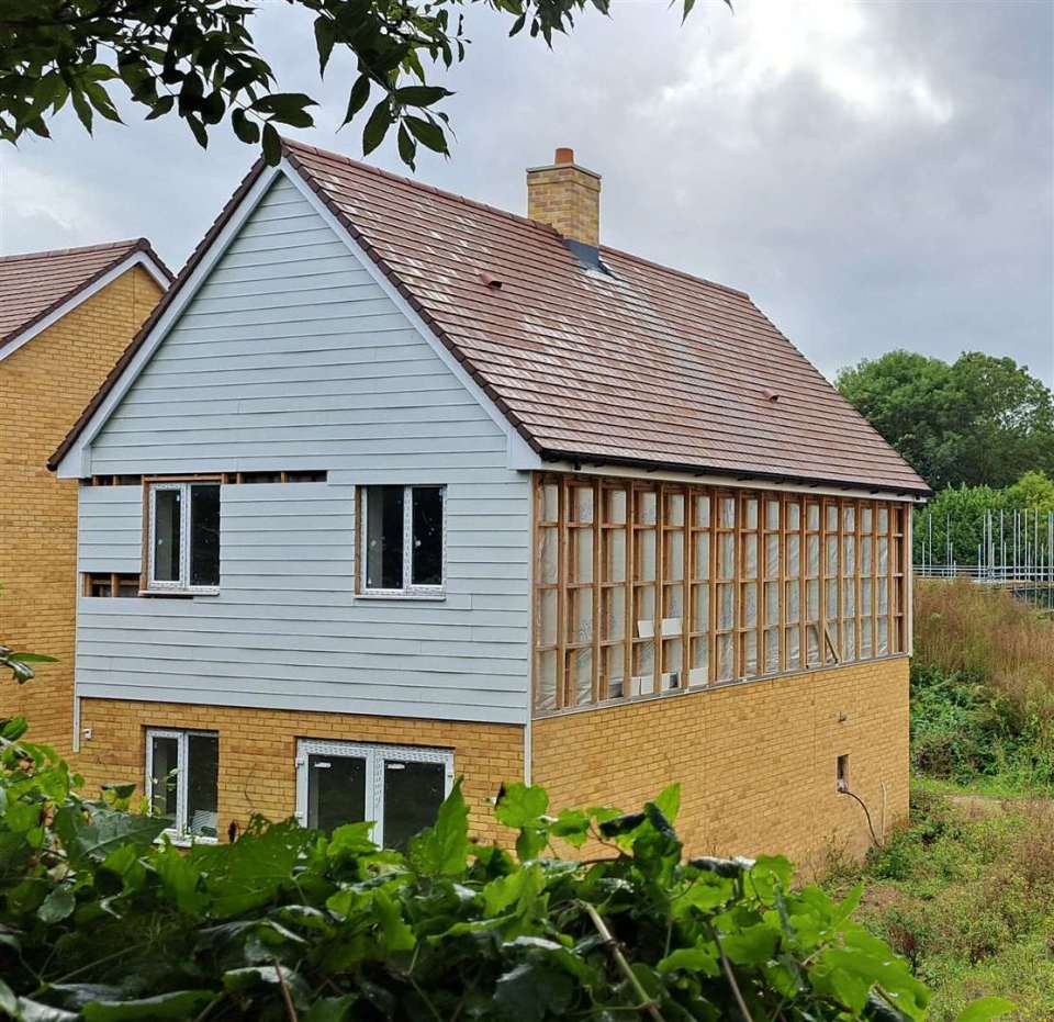 An unfinished house with a partial light grey facade, exposed wooden framework, and yellow brick, seen through green foliage.