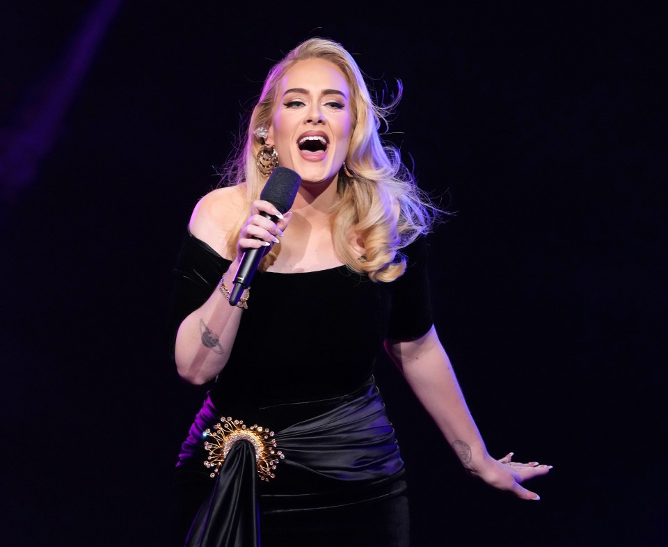 Adele performing onstage, wearing a black off-shoulder dress with a jewel-encrusted belt.