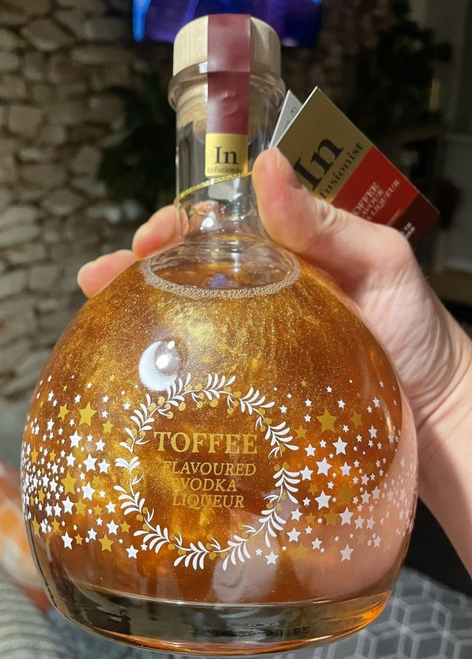 A hand holding a bottle of Aldi's Toffee Flavored Vodka Liqueur, which is a shimmery golden color with white and gold stars on the bottle.