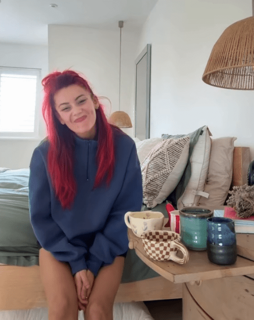 Woman with red hair sitting on a bed, smiling at the camera, with several mugs on a bedside table.