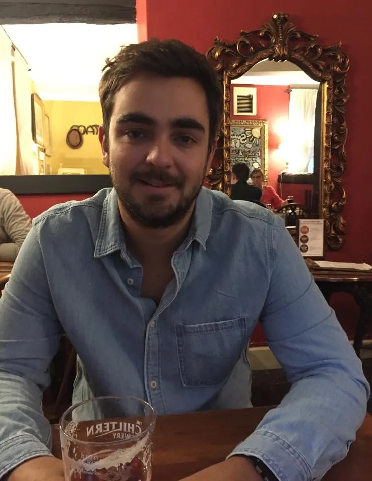 Nick Barnard, a man with a beard and dark hair, wearing a denim shirt, seated at a wooden table with a glass of beer.