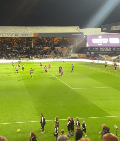 Football players warm up on a brightly lit field at night, while spectators watch from the stands.