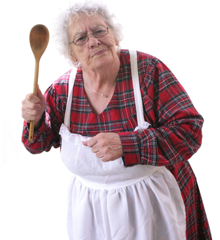 A senior woman in a red plaid shirt and apron holds a wooden spoon with a stern expression.