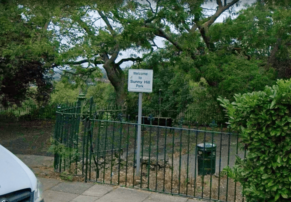 Sign reading "Welcome to Sunny Hill Park" behind a black metal fence, with lush green trees and foliage.