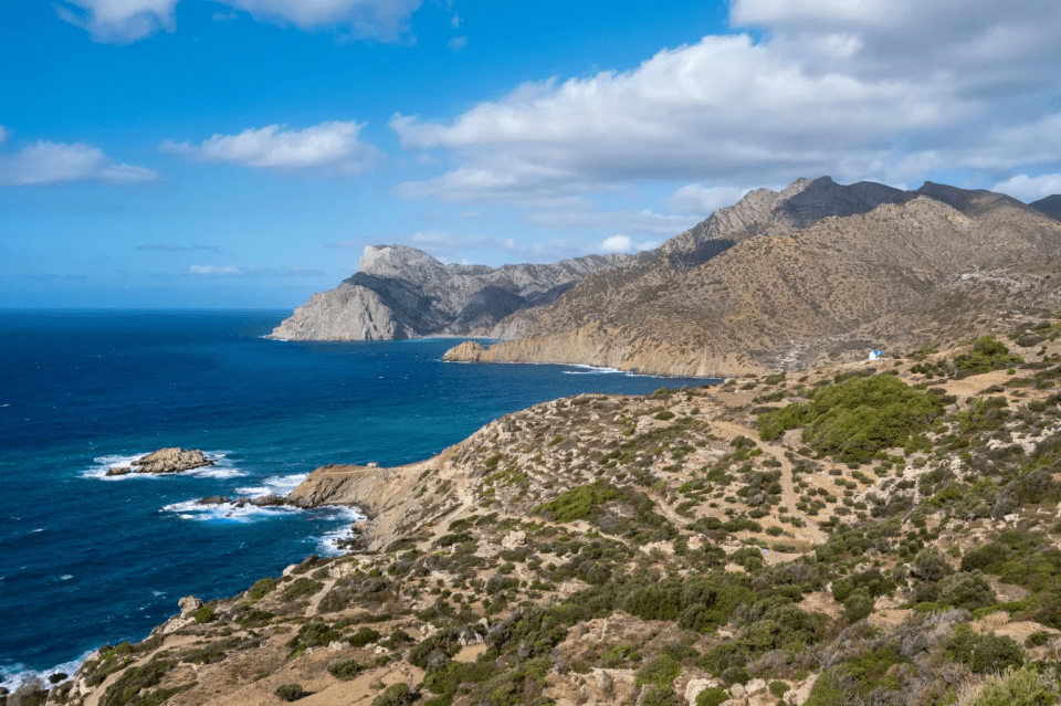 Coastal landscape with mountains, blue sea, and a small white building.
