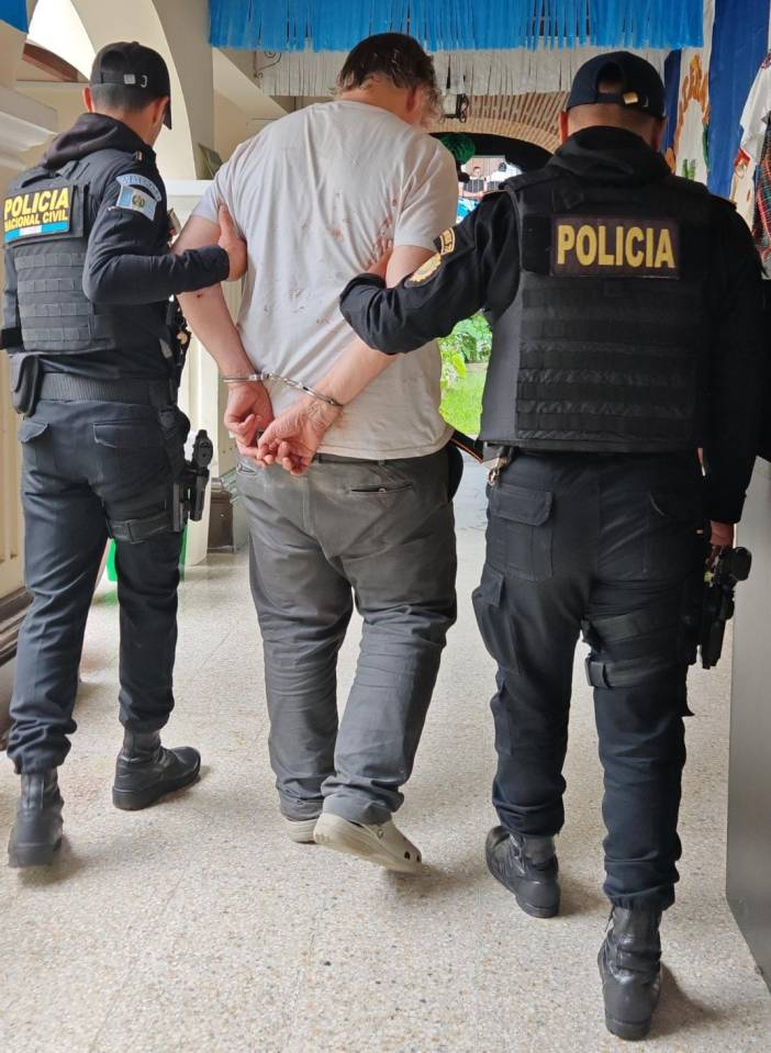 A man in handcuffs, wearing a white t-shirt stained with blood, is being escorted by two police officers in black tactical gear.
