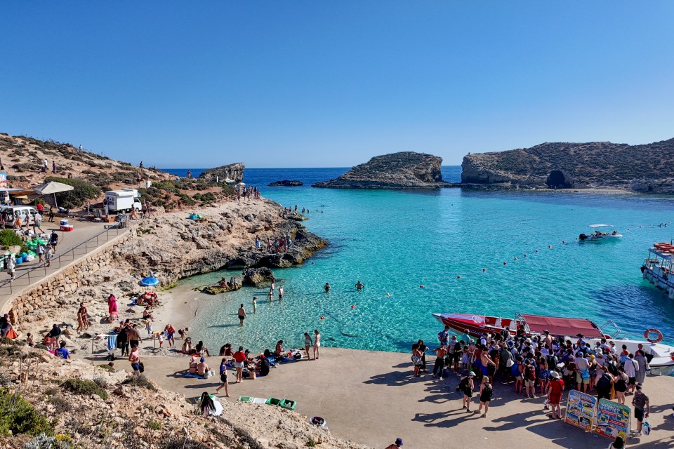 Aerial view of a bay in Blue Lagoon, Comino, Malta, bustling with people enjoying the beaches, swimming in the clear blue water, and waiting to board boats along the rugged coastline.
