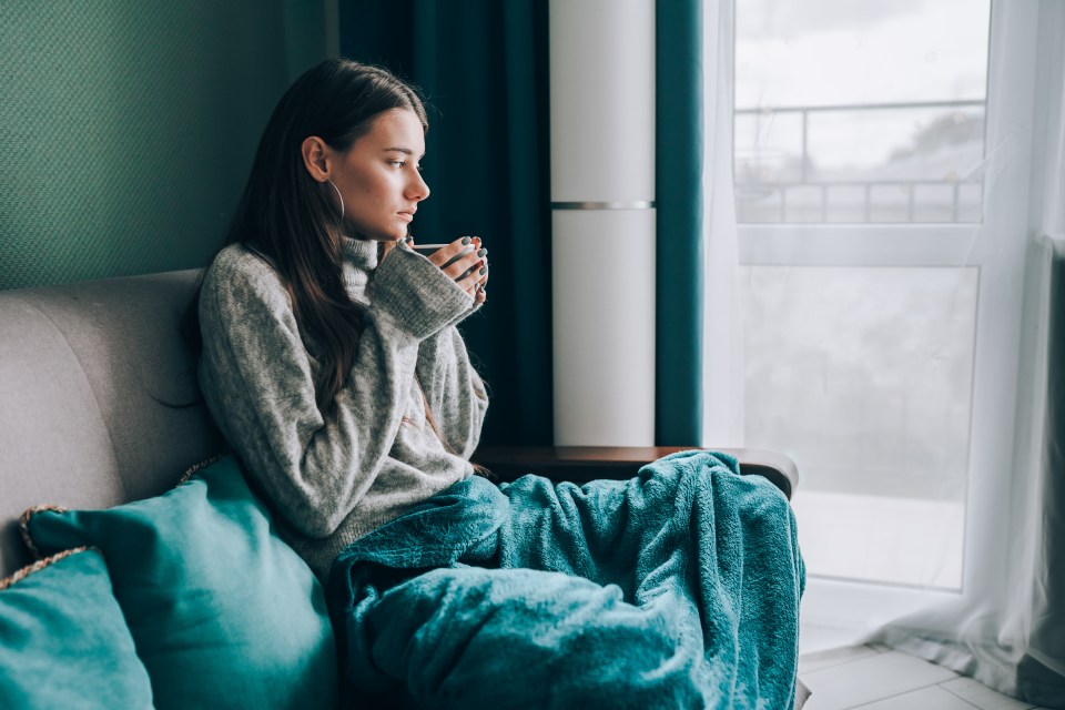 Woman sits on a sofa wrapped in a blanket, drinking from a mug.