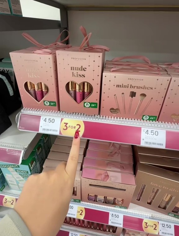 A person pointing at a "3 for 2" offer for "nude kiss" lip gloss and "mini brushes" sets priced at £4.50 each in a store aisle.