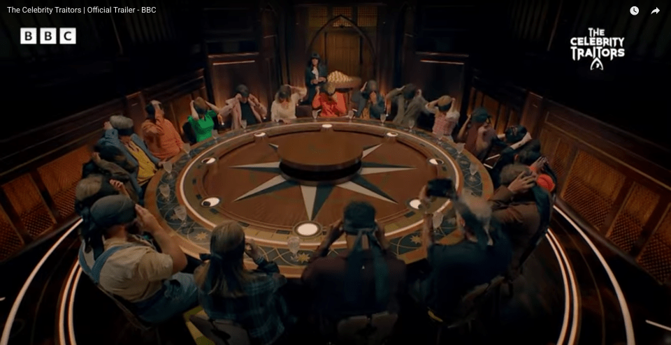 A circular table of contestants in a room with a moderator standing in the back.