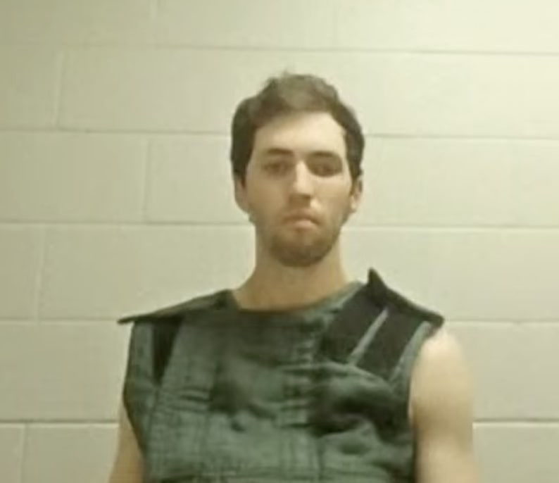 A mugshot of a man with short brown hair and a beard, wearing a dark colored vest.