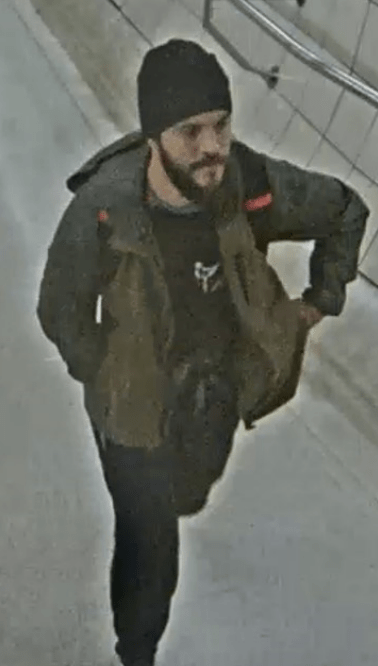 CCTV image of a man with a beard and dark beanie, wearing a dark jacket, walking down a tiled hallway.