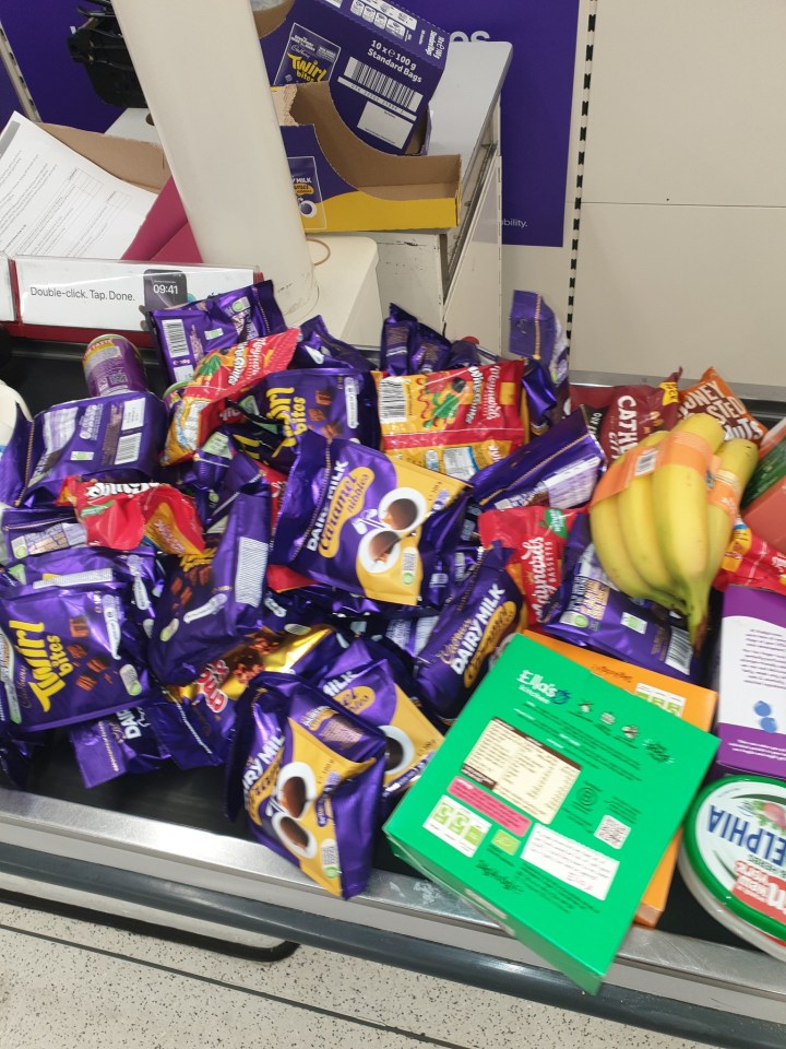 Shopping cart full of groceries, including bags of chocolates, bananas, and a container of Philadelphia cream cheese, on a supermarket checkout conveyor belt.