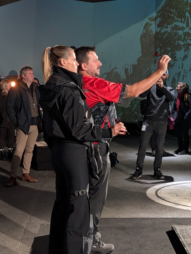 A woman in a black jacket and a man in a red and black shirt with a dart in his hand, looking at a screen projected with a mountainous image.