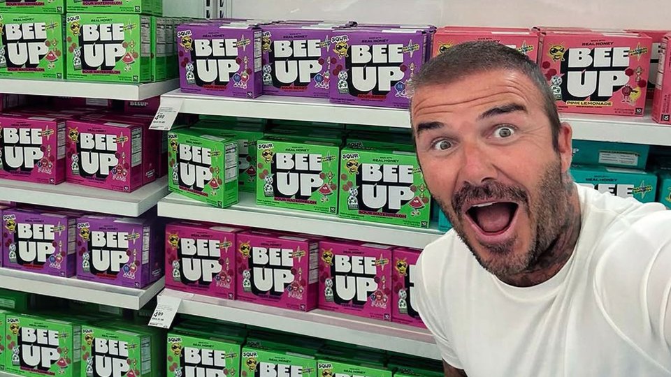 David Beckham smiling at the camera in a store aisle with multiple shelves of Bee Up energy drinks in various flavors.