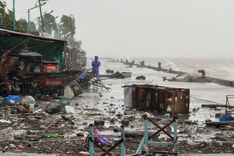 A man in a raincoat standing on a debris-strewn waterfront road with large waves crashing in the background due to Super Typhoon Ragasa.