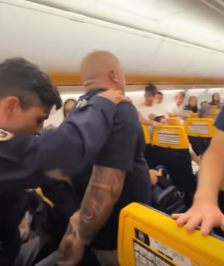 Two men in uniform detain a man on a crowded airplane.