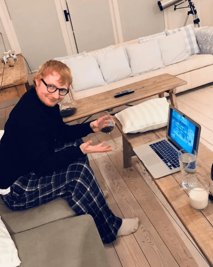 Ed Sheeran at home, holding a glass of wine, sitting on a couch with a laptop.