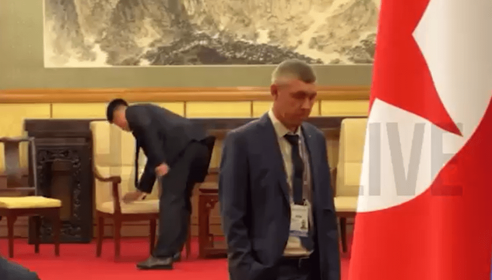 A man in a suit stands near a flag while another man arranges chairs.