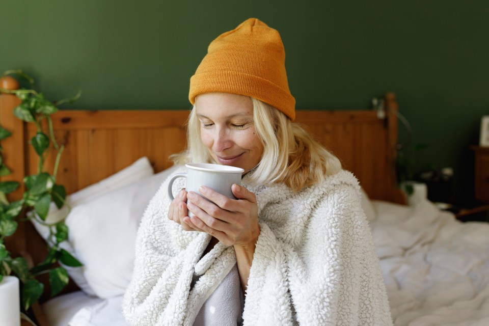 Smiling mature woman in a knit hat, wrapped in a blanket, holding a cup and drinking a hot beverage in bed.