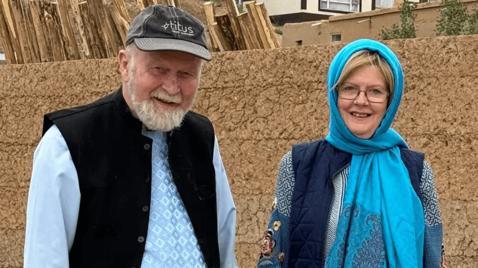 A man in a black vest and a woman in a blue headscarf smile at the camera.